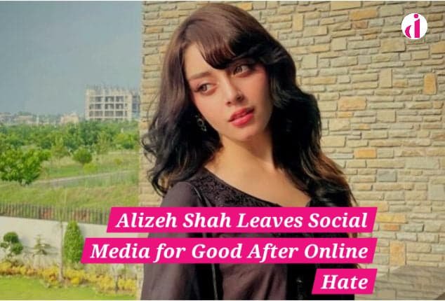 Alizeh Shah poses outdoors in a black outfit, standing near a stone wall with a distant look, amid greenery and buildings in the background.